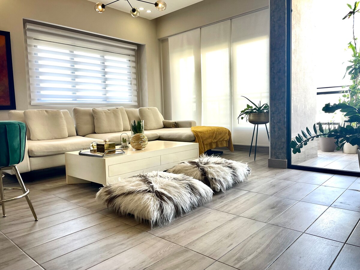 A spacious living area features a light-colored sectional sofa complemented by decorative pillows. A low coffee table sits in the center, adorned with books and a decorative plant. Soft rugs are placed on the tiled floor, enhancing the cozy atmosphere.