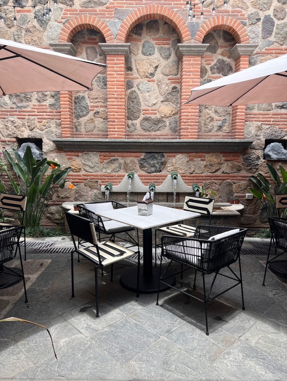 An outdoor dining area features a central round table surrounded by black metal chairs with cushions. Light-colored umbrellas offer shade, while a stone wall serves as a backdrop, decorated with arched niches and greenery. A water feature adds a serene element to the space.