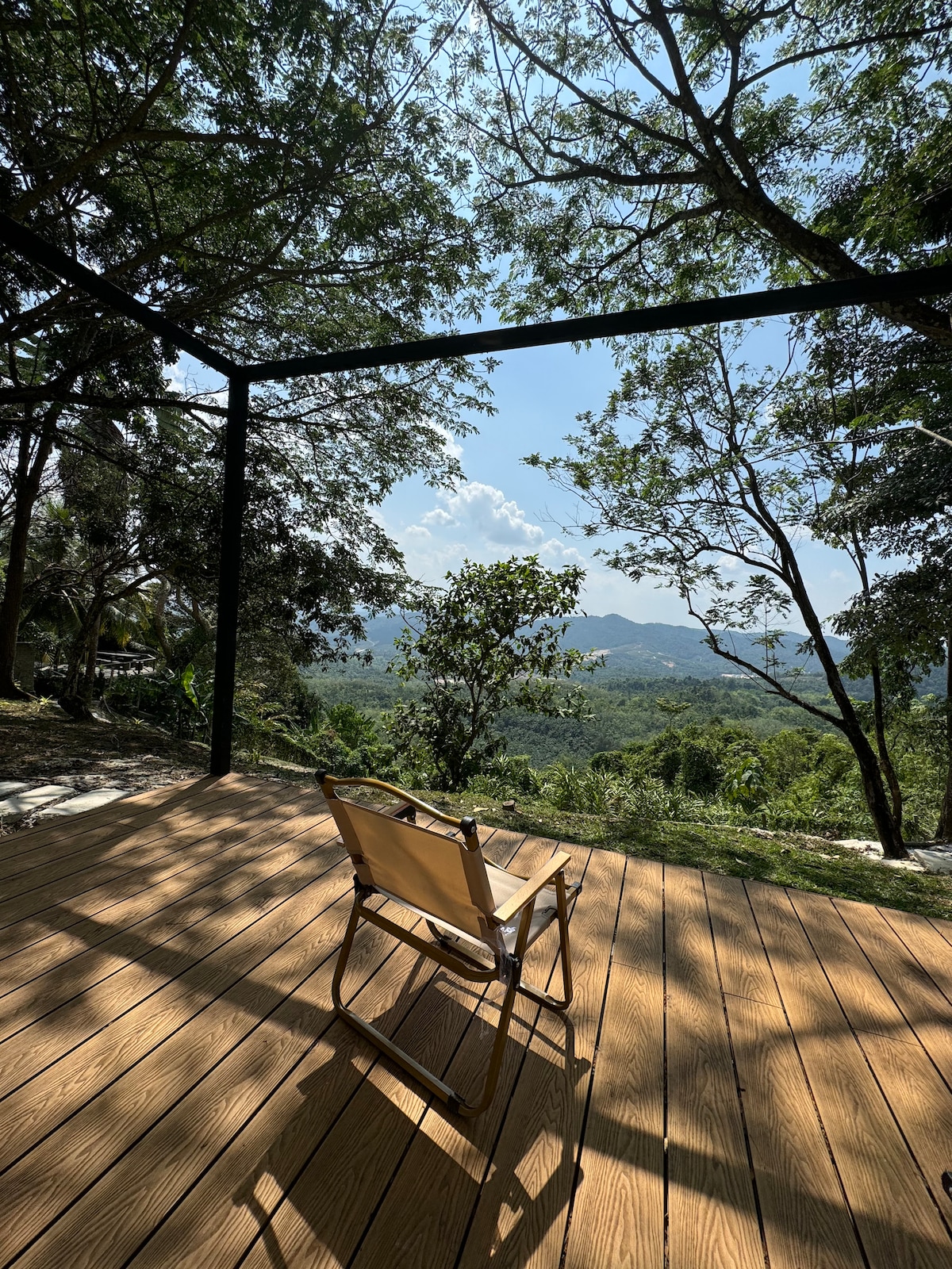 A spacious wooden deck features a single, lightweight chair positioned to face a panoramic view of the surrounding forest and distant hills. Lush greenery frames the scene, with sunlight filtering through the trees, creating a serene outdoor retreat.