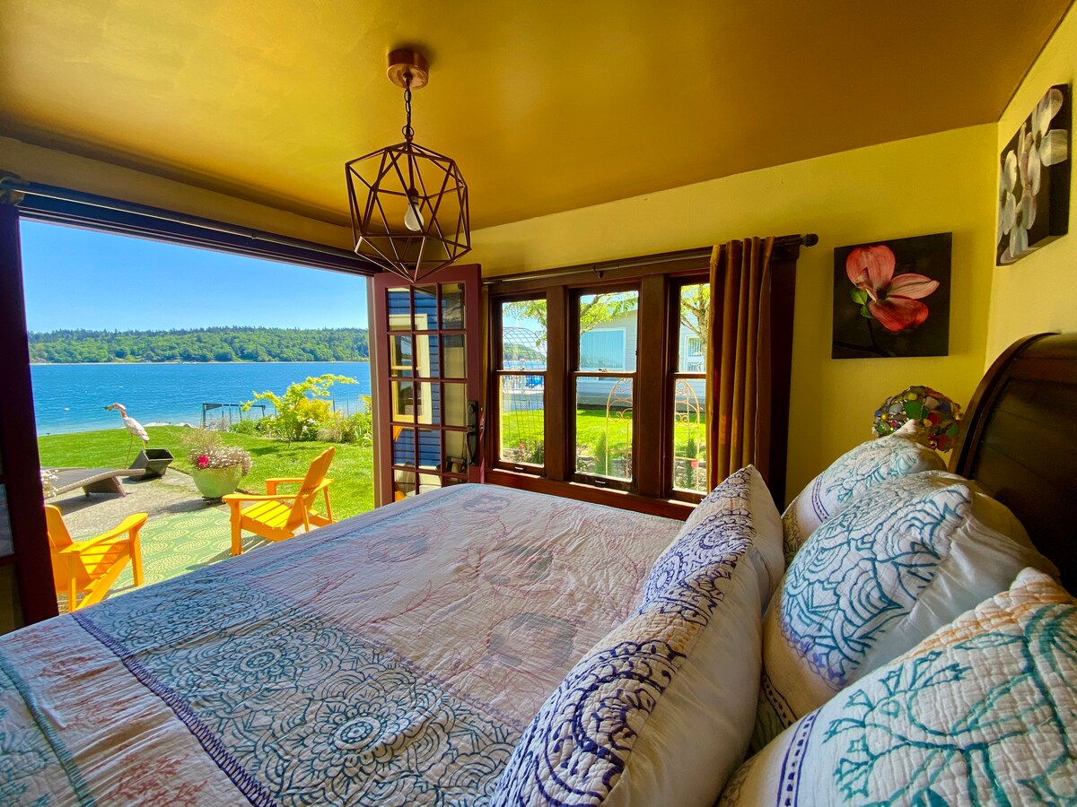 A master bedroom with a king size bed features vibrant pillows and a colorful bedspread. French doors open to a patio with outdoor seating and views of the tranquil harbor. Natural light illuminates the space, enhancing the warm yellow walls.