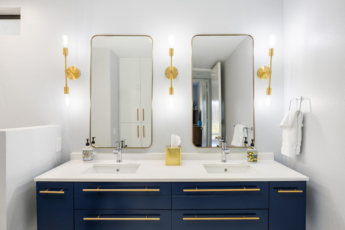A modern bathroom features a double sink vanity with a sleek white countertop and navy blue cabinetry. Two framed mirrors are placed above each sink, complemented by stylish wall sconces. A clean, bright ambiance is created by the white walls and ample light reflected throughout the space.