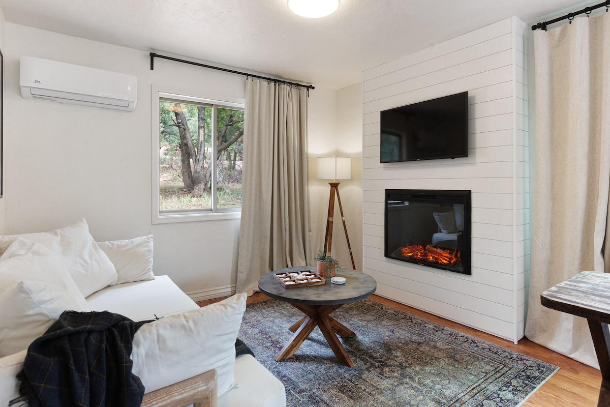 A comfortable living area features a modern TV mounted above a cozy electric fireplace. Soft, neutral curtains frame a window that allows natural light to enter. A round coffee table is centered on a patterned rug, with a plush couch inviting relaxation.