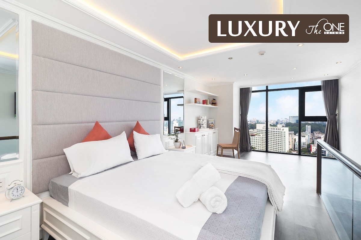 A bright bedroom featuring a spacious bed with soft linens and plush pillows. Large windows allow natural light to fill the room, offering a view of the surrounding cityscape. Minimalist decor creates an airy atmosphere, complemented by a few decorative items on nearby shelves.