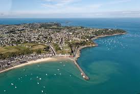 Aerial view showcases a coastal town surrounded by a sandy beach and calm turquoise waters. Small boats dot the harbor, while green landscapes and residential areas line the shore. The scene highlights the tranquil blend of nature and community, with a tranquil sky complementing the natural beauty.