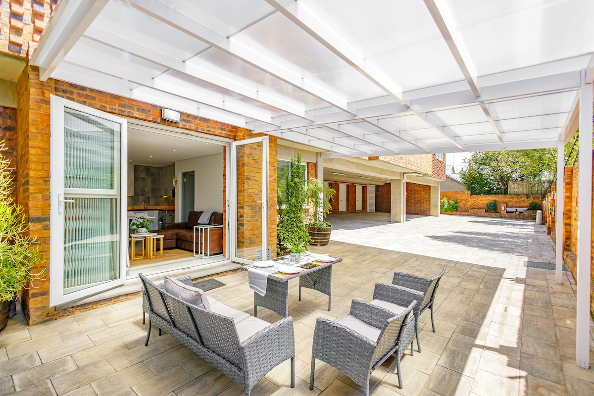An outdoor seating area is presented, featuring a compact table surrounded by four wicker chairs. A covered patio offers protection from the sun, while the surroundings consist of a spacious, paved area bordered by greenery and brick walls.