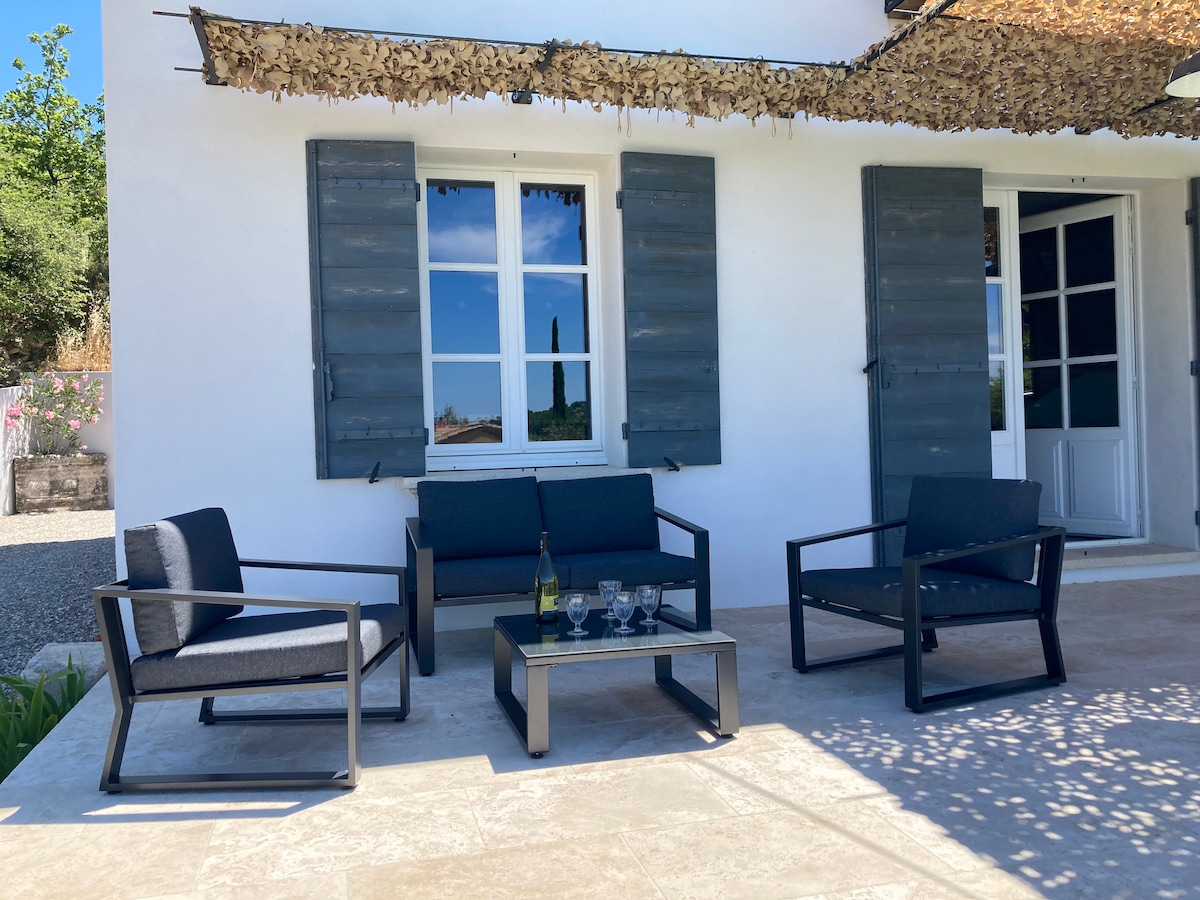 A small outdoor seating area is depicted, featuring two armchairs and a sofa facing a coffee table. The backdrop includes an exterior wall with large, open shutters and a door. Natural shade from above creates a comfortable spot for relaxation.