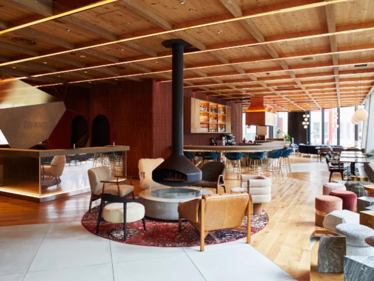 The lobby area features a large open design with wooden ceilings and a modern fireplace at the center. Various seating options are arranged around a circular rug, complemented by a stylish bar area lined with high stools and warm lighting.