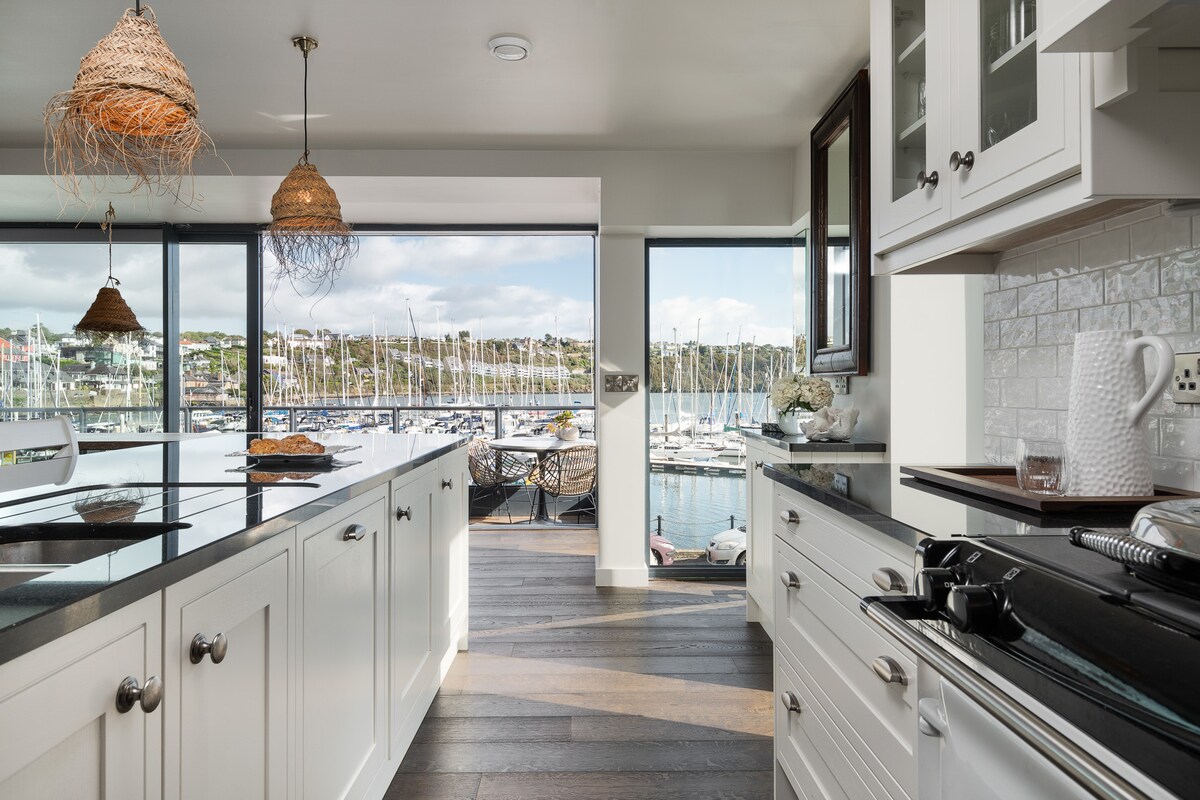 A modern kitchen is presented with white cabinetry and sleek black countertops. Large windows frame a view of the marina, allowing natural light to enhance the open space. Unique lighting fixtures hang from the ceiling, adding a stylish touch to the functional area.
