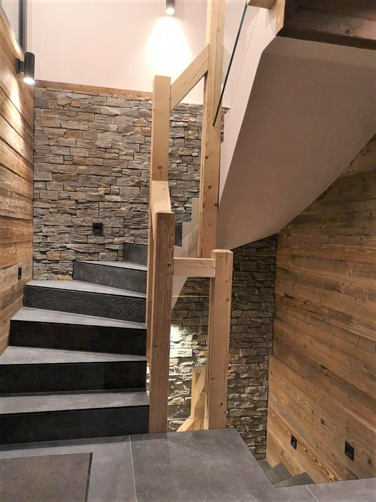 A staircase features a combination of natural wood and stone elements, creating a rustic yet modern aesthetic. Soft lighting illuminates the steps, while a glass railing offers a sense of openness. The walls are adorned with wooden panels, enhancing the inviting atmosphere.