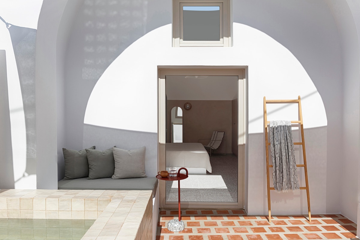 A shaded terrace area features a sleek, shallow pool beside a comfortable seating space with gray cushions. A ladder holds a soft towel, and a stylish table holds a drink, with an inviting indoor room visible through the open glass door.