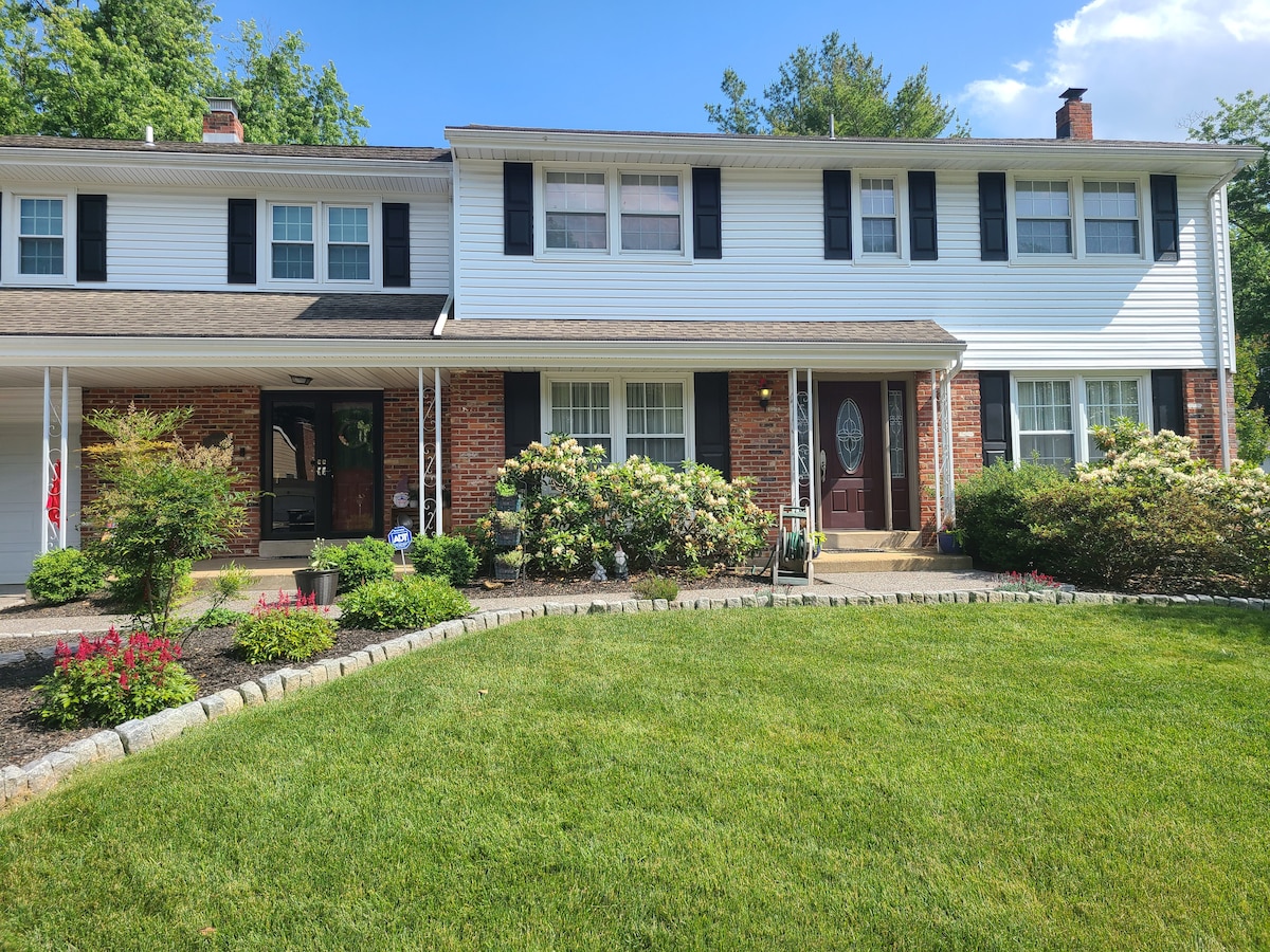 The front exterior of a two-story home is presented, featuring a combination of brick and siding. The neat lawn is bordered by a stone walkway, and flowering shrubs add color alongside the front entrance. Large windows provide natural light to the interior.