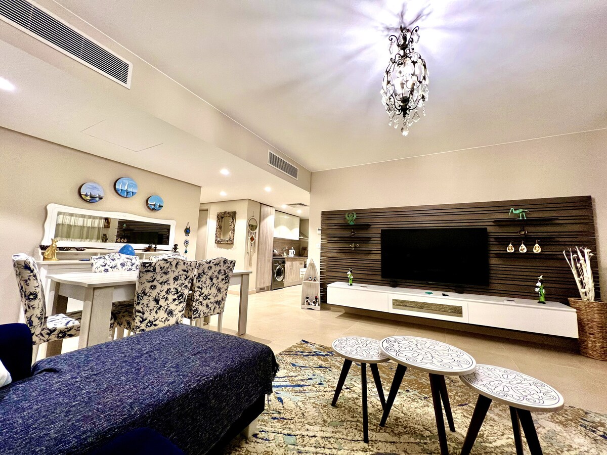 A spacious living area features contemporary furnishings and a large wall-mounted television. Elegant light fixtures hang from the ceiling, while a soft area rug complements the color palette. A dining space with floral-patterned chairs is visible, contributing to an inviting atmosphere.
