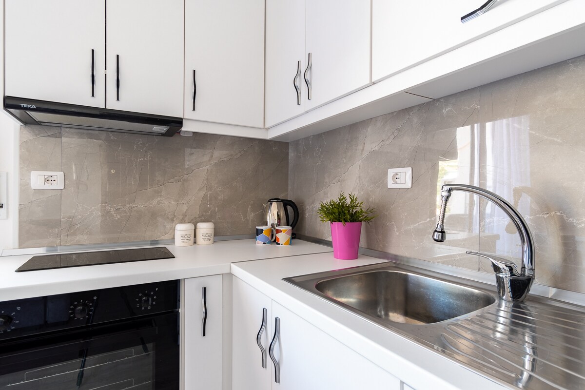 A modern kitchen features sleek white cabinetry and a polished light grey stone backsplash. An integrated electric stove and a preparation area are visible. A kettle and kitchenware sit nearby, accompanied by a small potted plant in a pink pot, adding a touch of greenery.