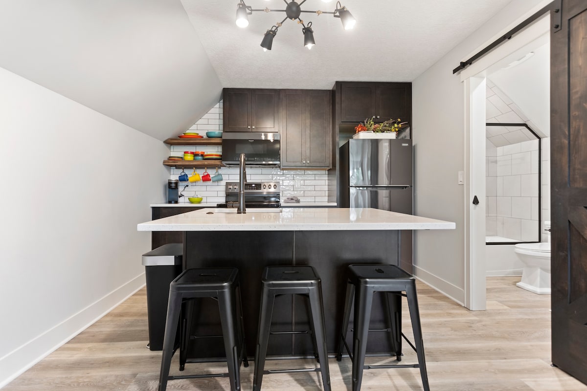 A modern kitchen features dark cabinetry and a white countertop, complemented by open shelving displaying colorful kitchenware. Three black bar stools line the kitchen island, with stainless steel appliances and stylish light fixtures visible above. The doorway on the right leads to the bathroom.