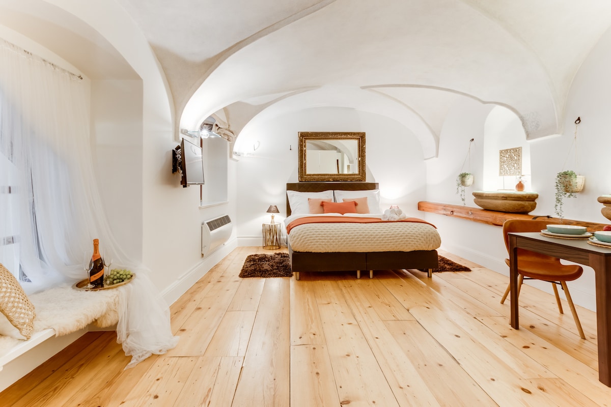 A spacious studio features an elevated ceiling with elegant archways. A king-size bed, adorned with neutral bedding, is complemented by a large mirror and subtle lighting. A wooden table with chairs and dishes is present, alongside a cozy seating area with natural light filtering through rounded windows.