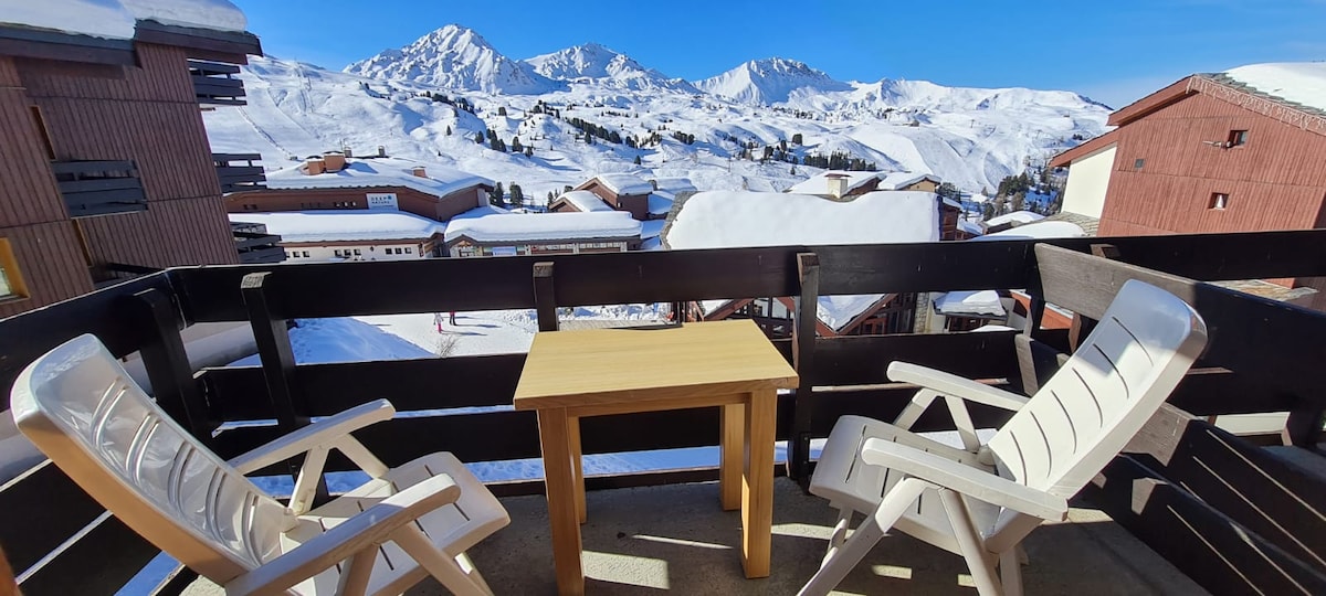 A balcony is featured with two white chairs and a small wooden table, providing a view of snow-covered mountains and slopes. Surrounding buildings are also visible, with clear blue skies above enhancing the tranquil setting.