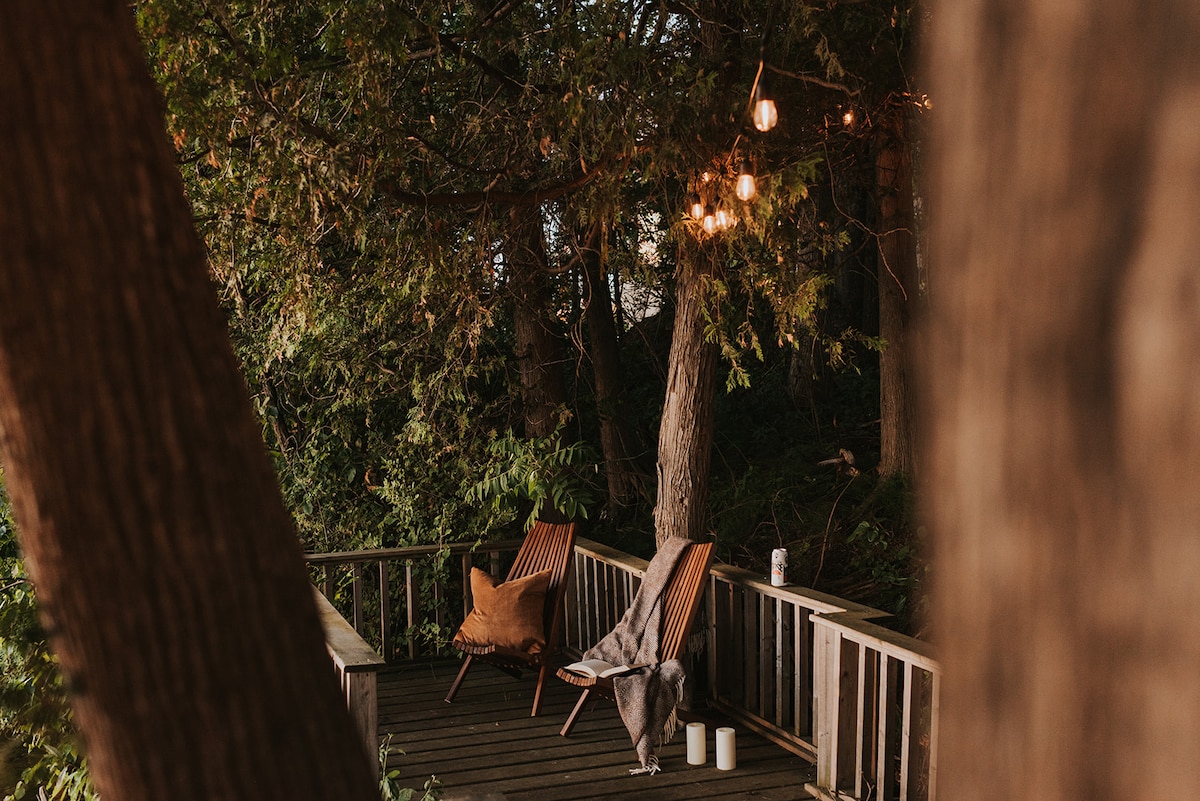 A serene lakeside patio features two wooden chairs, one draped with a soft blanket, positioned along a wooden deck surrounded by trees. Soft lights hang above, creating a calming atmosphere for relaxation.