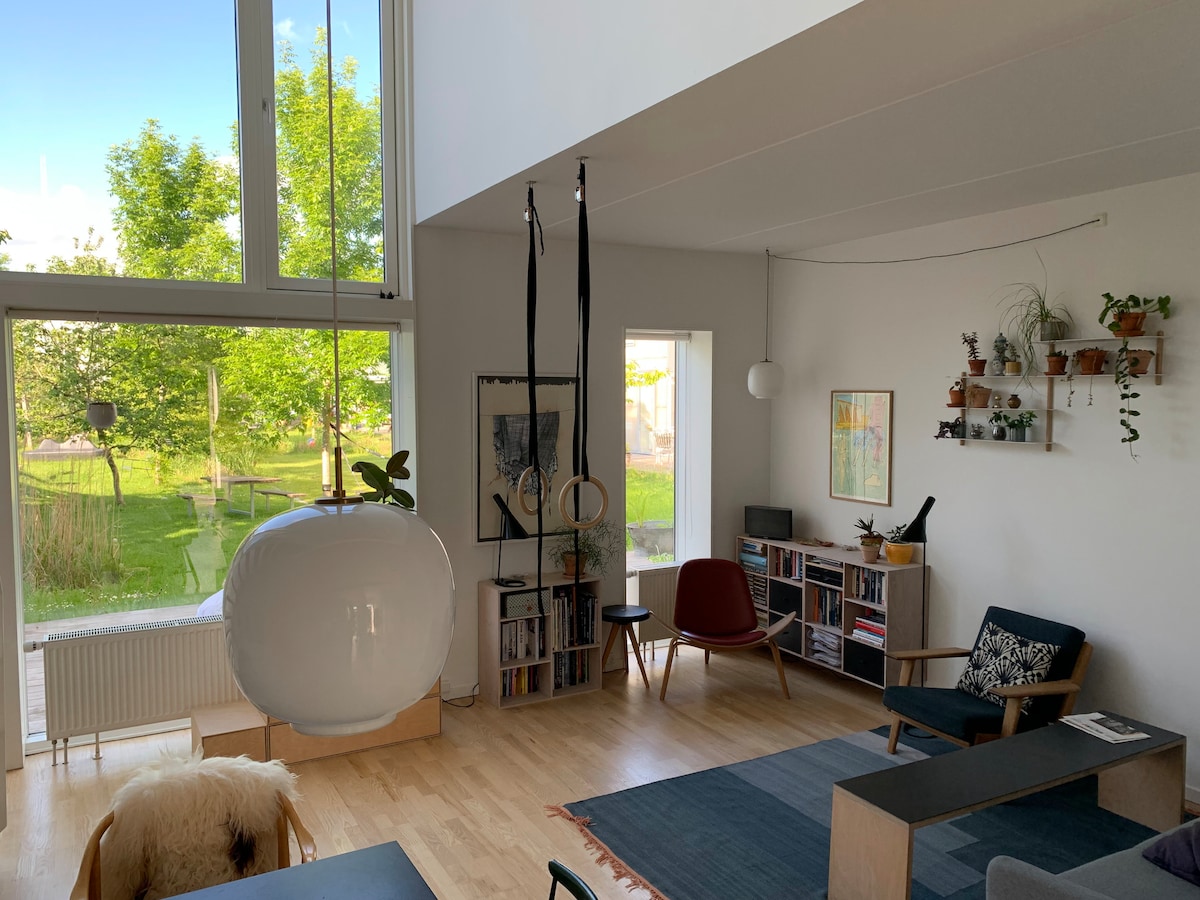 A bright living area features large windows that bring in natural light and frame views of greenery. A modern pendant light hangs above a minimalist coffee table. Comfortable seating options include wooden chairs and a classic armchair, complemented by a soft area rug.