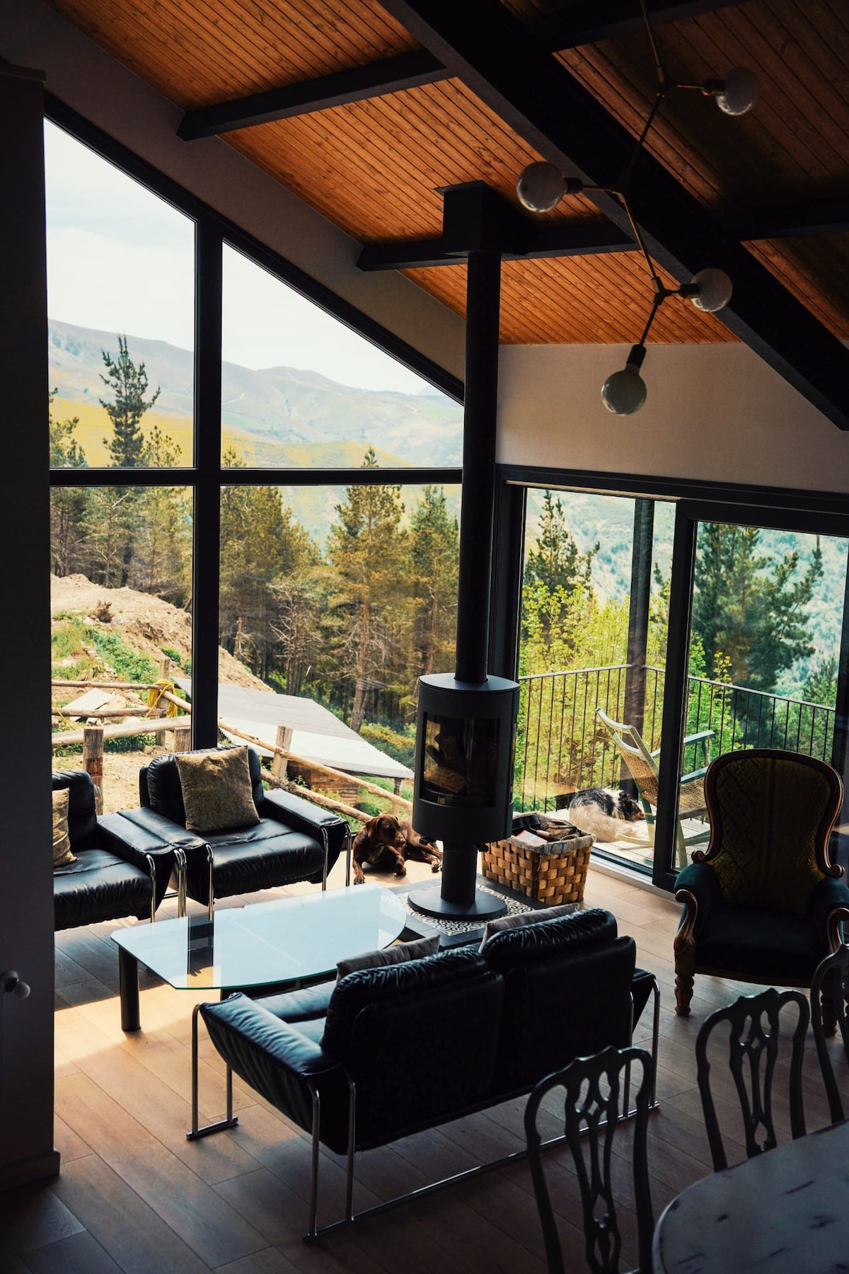 A spacious living area offers floor-to-ceiling windows that frame mountain views and surrounding trees. Comfortable black seating is arranged around a sleek glass table. A modern fireplace adds a cozy touch, while a balcony extends the space outdoors, enhancing the connection to nature.