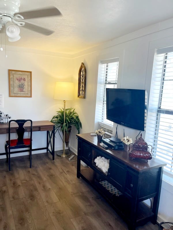 A comfortable living area features a wooden entertainment console with decorative items. A flat-screen TV is positioned on top, and a desk with two chairs sits nearby, illuminated by a standing lamp. Natural light filters through window blinds, enhancing the welcoming atmosphere.