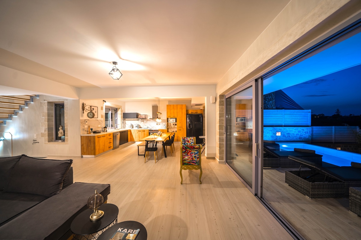 An open-plan living area features large sliding glass doors that provide a view of the outdoor space, softly illuminated by nighttime lighting. The room showcases a comfortable dark couch, a uniquely patterned chair, and a kitchen area with wooden cabinetry.