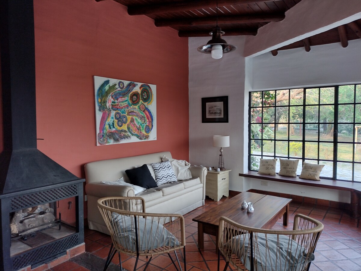 A cozy living area is highlighted by a white sofa and a wooden coffee table, positioned near a large window allowing natural light to fill the space. A modern fireplace and decorative art piece add character to the room, while comfortable seating is provided by wicker chairs.