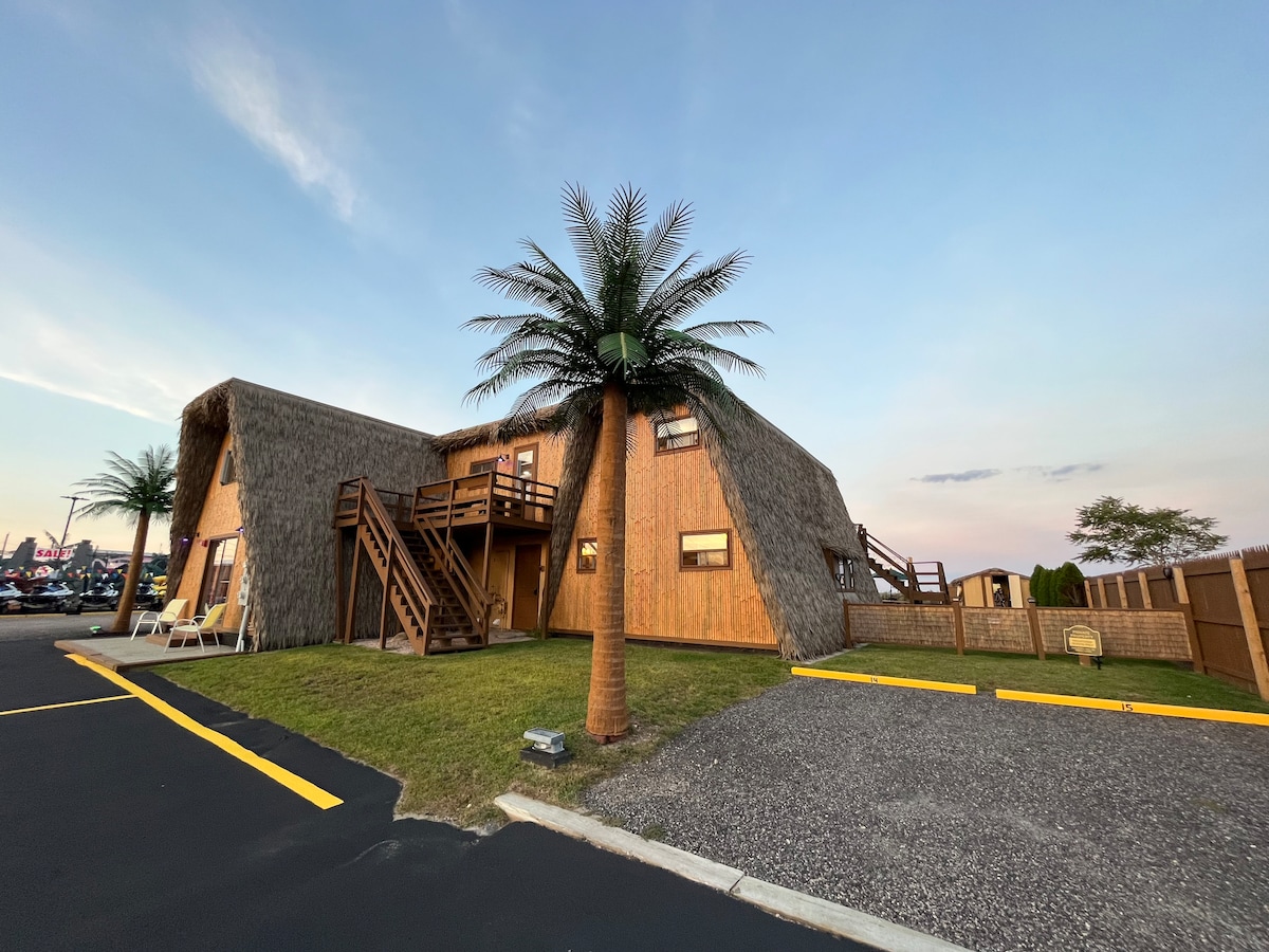 A unique two-story structure with a thatched exterior is depicted. The building features a wooden staircase leading to the second floor. Palm trees are positioned in the foreground, while a grassy area is adjacent to the building. A paved parking area is visible in the foreground.