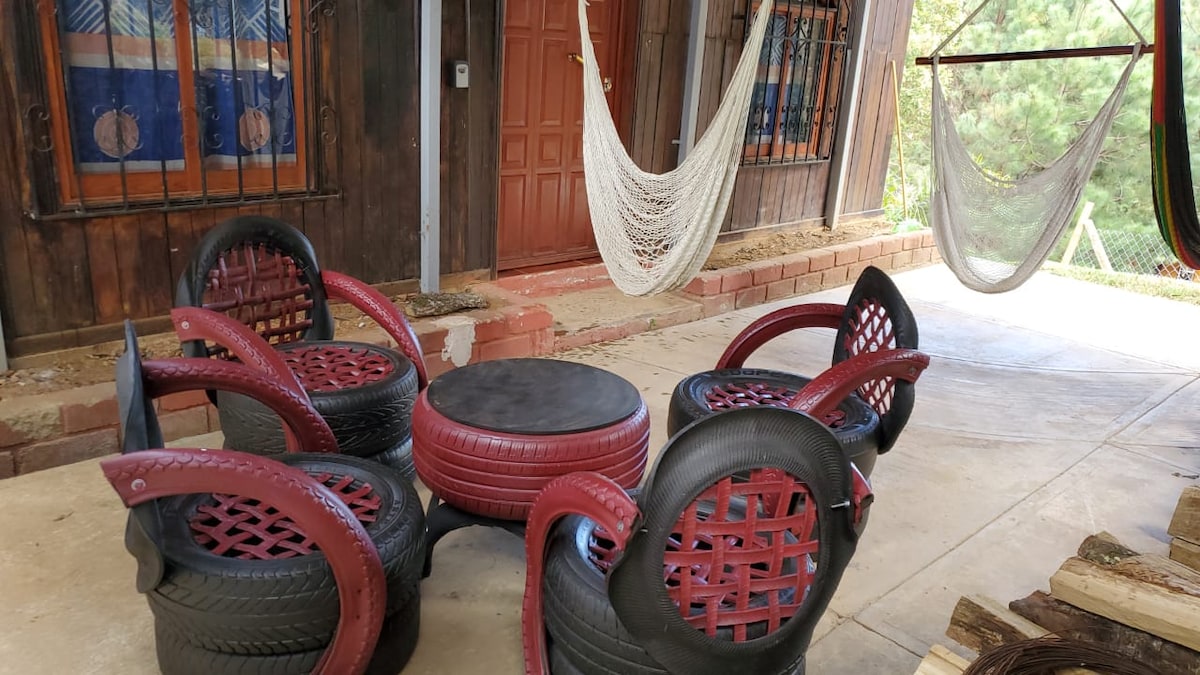 A covered porch area features six seating chairs made from repurposed tires, arranged around a circular table. Two hammocks hang nearby, inviting relaxation. Natural light filters through nearby windows, illuminating the rustic wooden walls of the cabin.