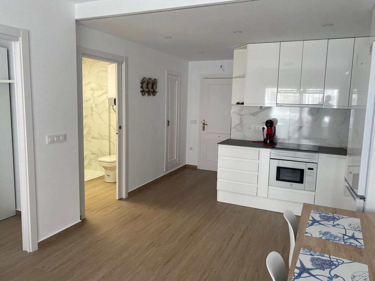 A modern kitchen is equipped with white cabinetry and a compact dining area. Natural light enhances the space, revealing clean lines and a neutral color palette. A small table is set with a patterned placemat, and appliances include a coffee maker and microwave.