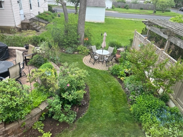 A lush garden view showcases a circular stone patio surrounded by a variety of greenery. Several patio chairs and a table provide seating, while tall plants and flowering bushes create a natural border. A wooden trellis adds structure to this serene outdoor space.