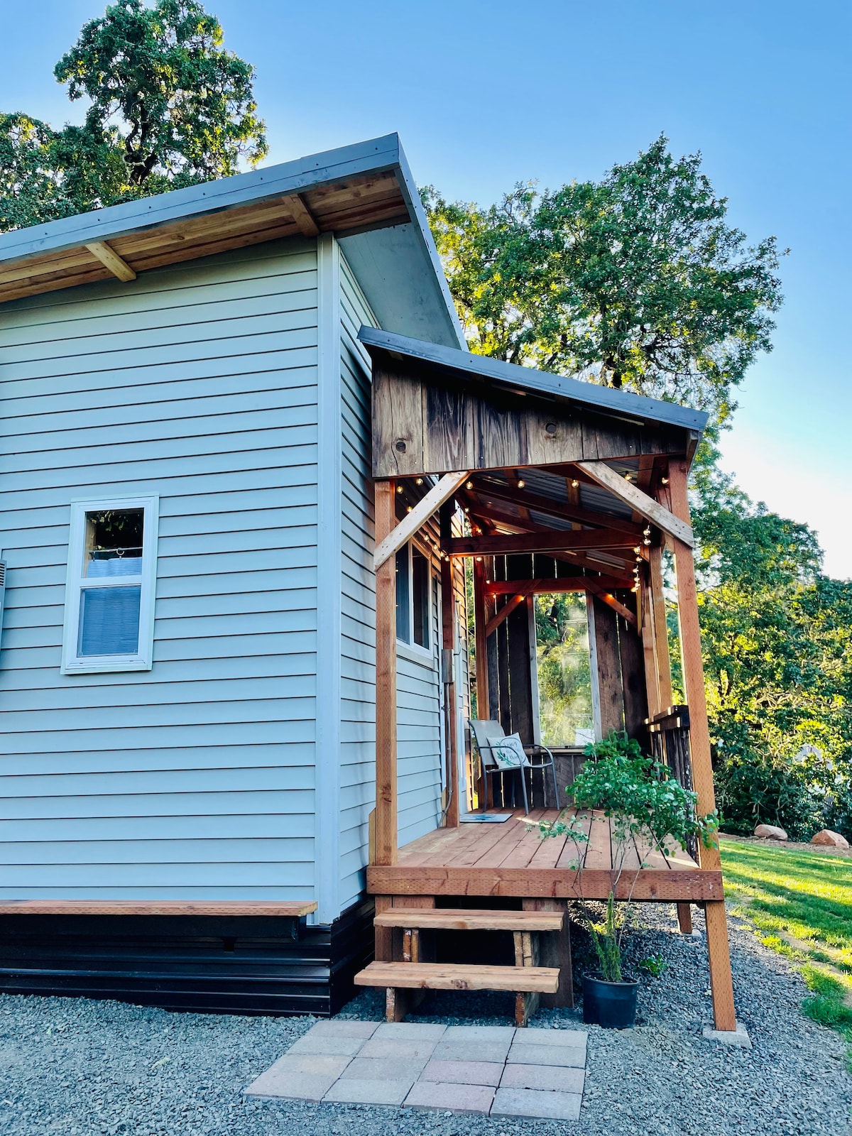 An outdoor porch is featured at the entrance of the home, constructed with wooden steps leading to a covered area. Two chairs are positioned on the porch, with sunlight filtering through the trees nearby, creating a serene environment.