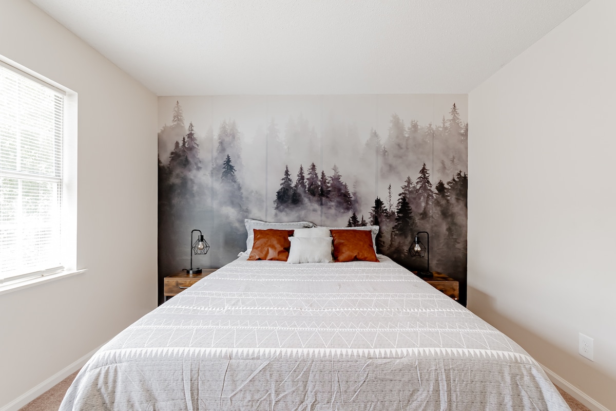 A spacious bed is positioned against a striking mural depicting foggy trees. Two decorative pillows in warm tones complement the neutral bedding. Bedside lamps provide soft lighting, enhancing the serene ambience of the sleeping area, with natural light filtering through the window.