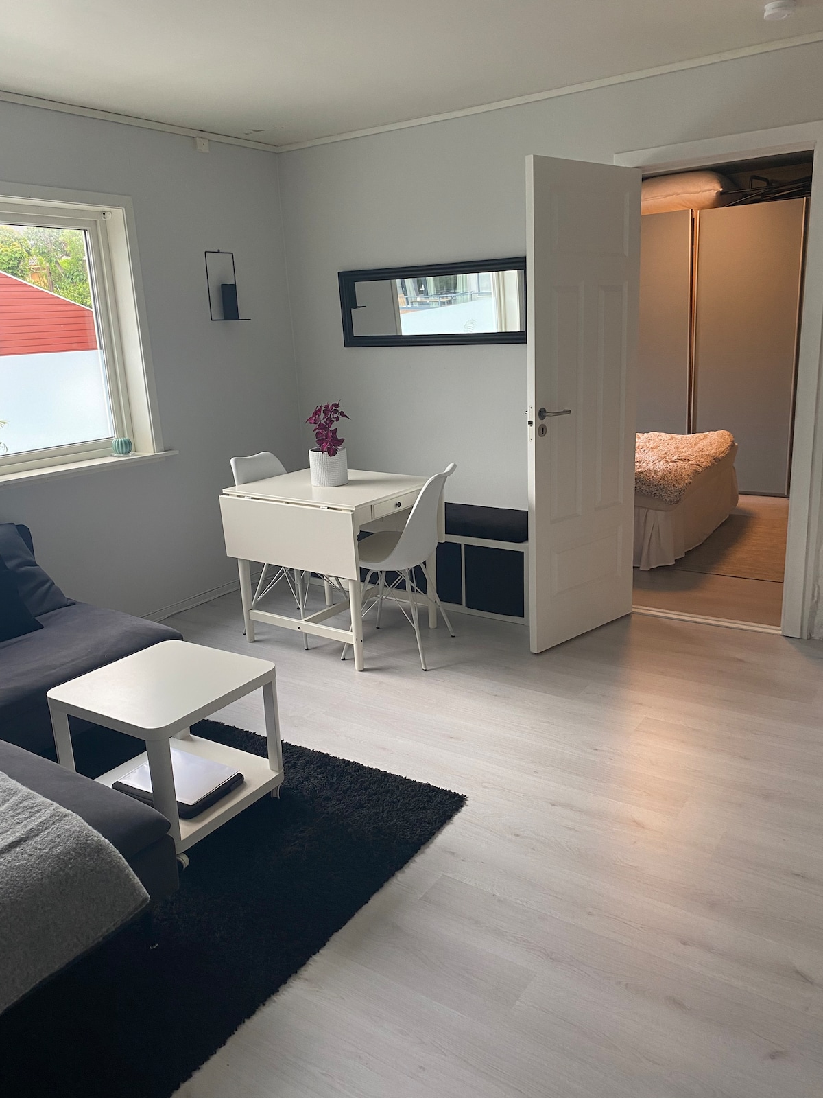 A light-filled living space features a dark area rug anchoring a seating arrangement with a gray sofa and a white side table. A small white desk with a plant and a mirror is visible, along with a doorway leading to a bedroom area, offering a serene layout.