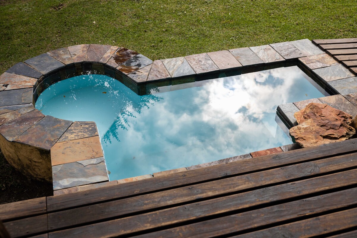 A small, oval-shaped swimming pool is set within a stone border, reflecting the sky above. Surrounding wooden decking offers a spacious area for relaxation. The pool's clear blue water invites leisure, while the grassy area beyond provides a natural backdrop.