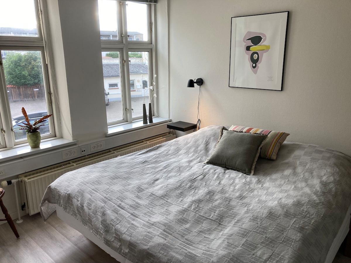 A spacious sleeping area features a large bed with a soft, patterned coverlet. A decorative pillow adds a touch of color. Sunlight filters through multiple windows, illuminating the room, while a modern artwork hangs above the bedside table.