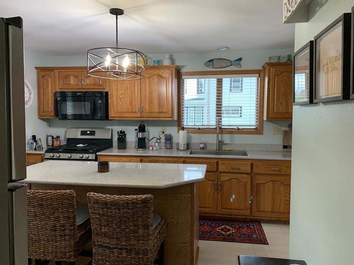 A well-equipped kitchen features wooden cabinetry and a large island with seating. A gas stove and microwave are visible beside the sink, under a window that allows natural light. The space is brightened by a modern, circular light fixture hanging overhead.