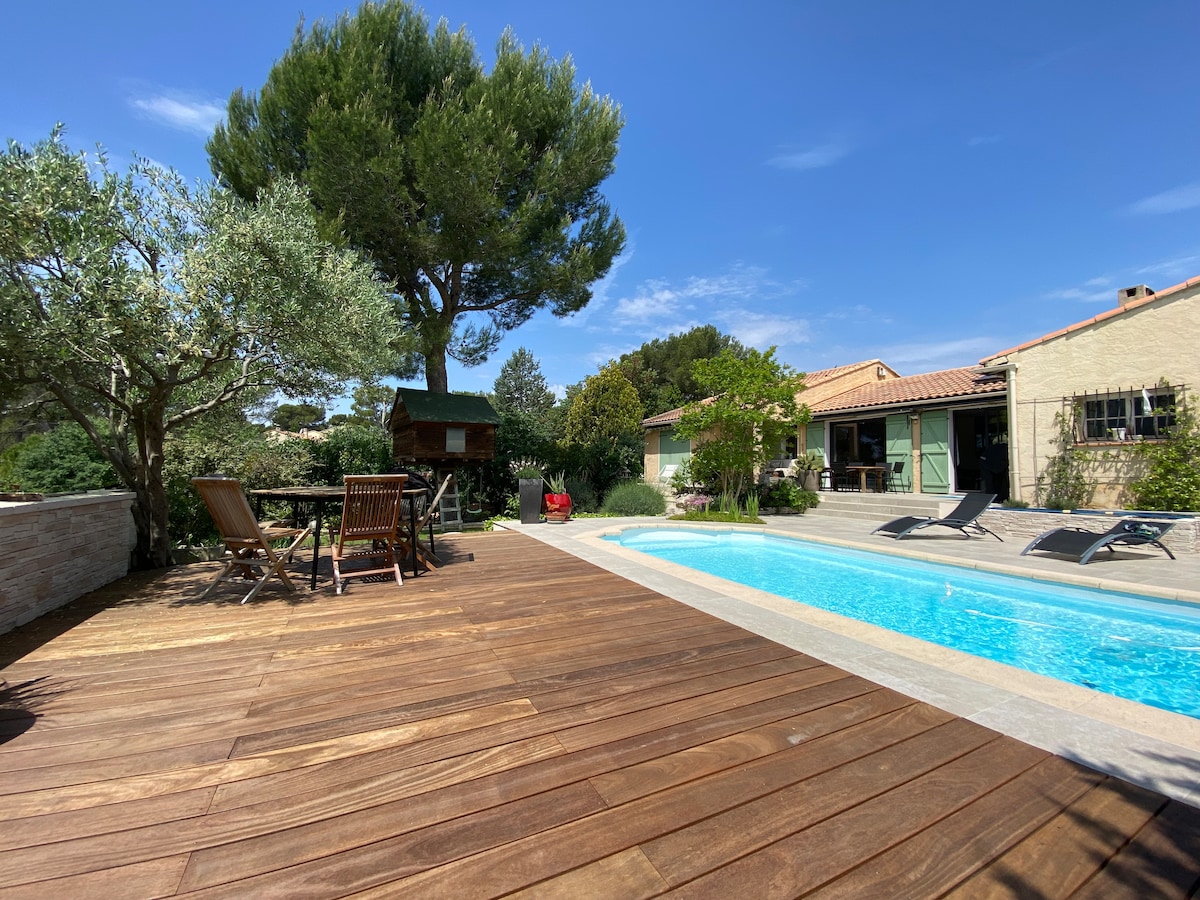 A wooden deck is featured alongside a private swimming pool, surrounded by greenery. Lounge chairs are positioned beside the water, while a small dining table with wooden chairs offers a space for relaxation. The house is visible in the background, nestled among trees.