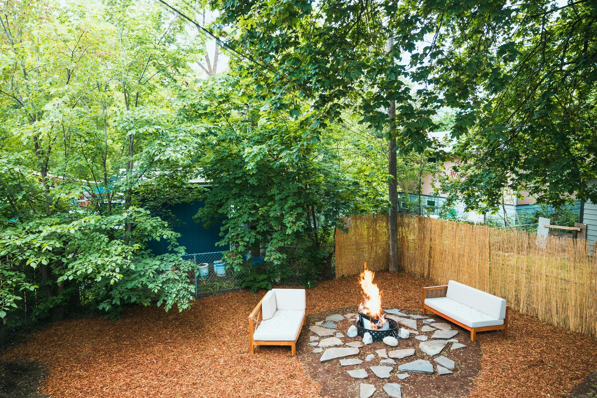 A shaded backyard features natural wood seating arranged around a stone fire pit, with a small fire glowing at the center. The area is surrounded by lush greenery and a bamboo fence, providing a tranquil space for outdoor gatherings.
