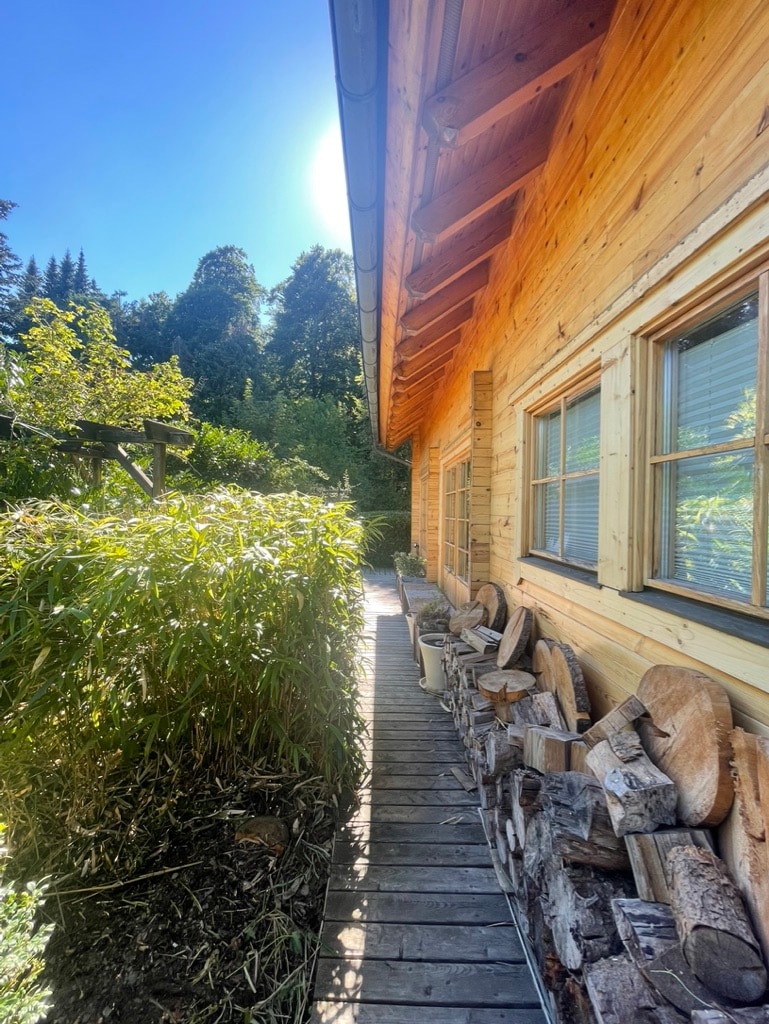The image shows a wooden pathway alongside a chalet, flanked by lush greenery and stacks of firewood. Two windows are visible, allowing natural light to brighten the space. The sun casts a gentle light over the scene, enhancing the tranquil outdoor environment.