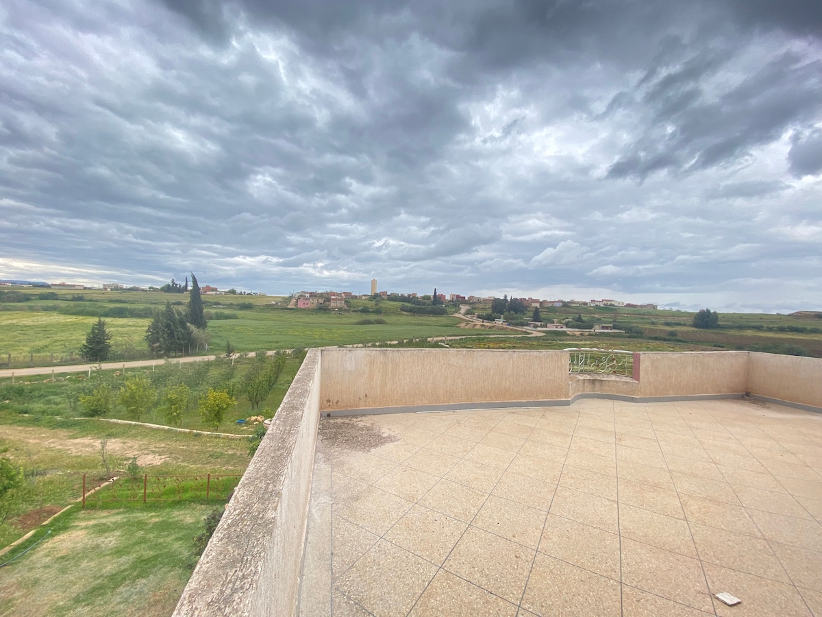 A spacious terrace offers panoramic views of expansive green fields under a textured cloudy sky. The outdoor area features a tiled floor extending towards the horizon, with landscape elements visible in the distance, creating an open and airy environment.