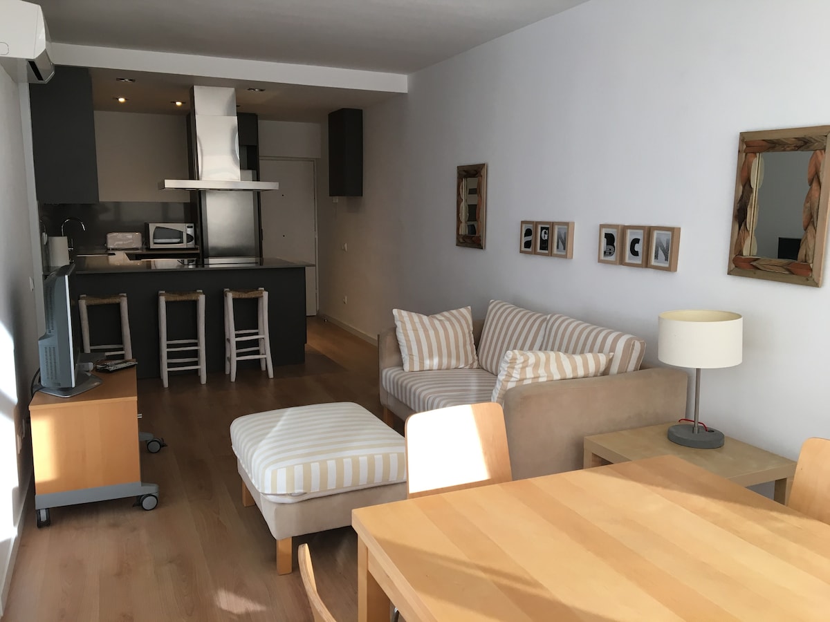 An open-plan living area showcases a neutral color palette, with a comfortable sofa featuring striped cushions and a matching ottoman. A wooden dining table is accompanied by light-colored chairs. The kitchen area includes a breakfast bar with stools, providing a modern and functional space.