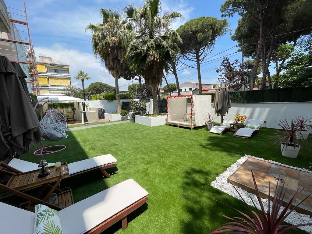A landscaped outdoor area is presented, featuring a lush green lawn and several sun loungers. Palm trees and decorative plants add to the ambiance. A shaded seating area and a wooden table are visible, creating a relaxing environment for guests to enjoy.