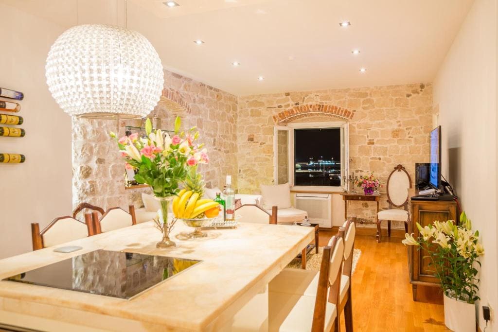 A dining area features a large table surrounded by wooden chairs, complemented by a stylish chandelier. The stone wall adds character, while a window provides a view of the outside. Fresh flowers and fruit adorn the table, enhancing the inviting ambiance.