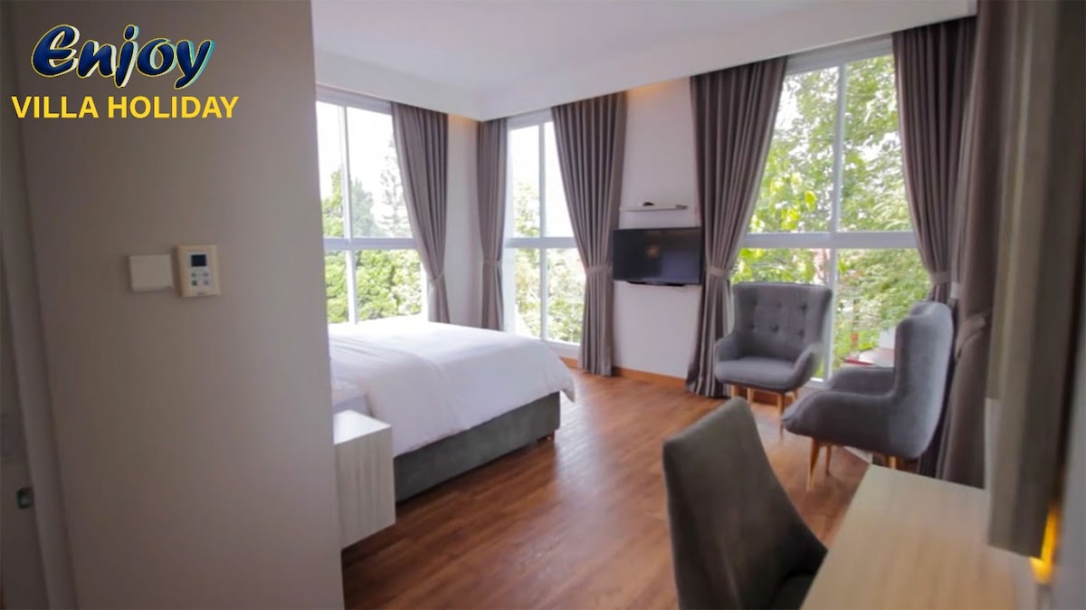 The interior of the room features large windows allowing ample natural light. A spacious bed is positioned against a wall, complemented by two armchairs and a desk. Soft curtains frame the windows, and a television is mounted nearby.