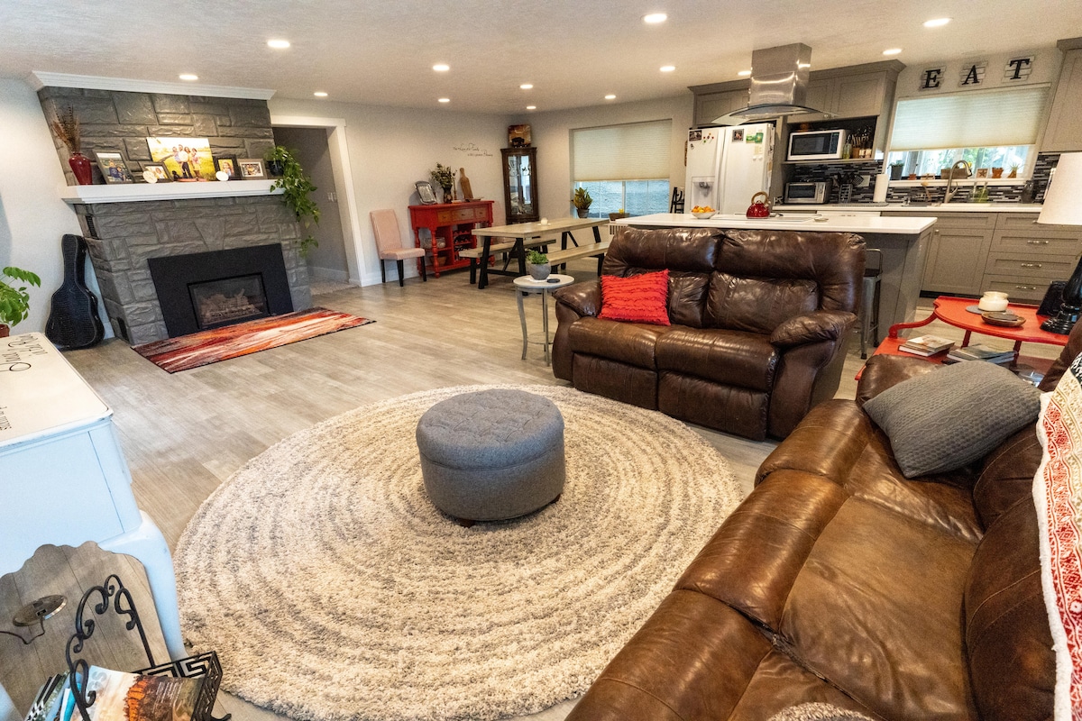 A spacious living area is highlighted by comfortable leather seating, including a large sectional sofa and a round ottoman. A circular area rug anchors the seating space, while a stone fireplace adds a cozy touch. The open layout leads into a well-equipped kitchen and dining area.