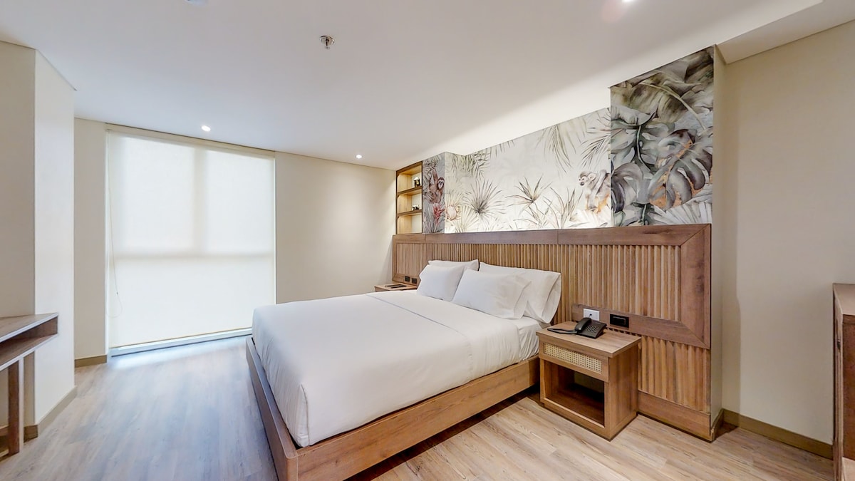 A light-filled bedroom features a spacious bed with crisp white linens against a vibrant mural of tropical plants. Wooden accents frame the bed and a side table, while a soft area rug enhances the warm wooden flooring. Large windows allow natural light to brighten the space.