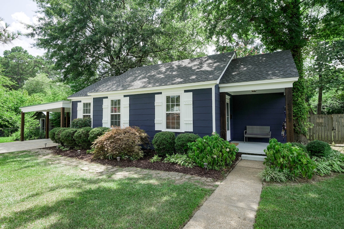 A cottage with dark blue siding is framed by neatly trimmed bushes and green grass. A covered porch features a bench alongside the main entrance, while tall trees provide shade in the background. A gravel driveway leads up to the home, adding to its inviting curb appeal.