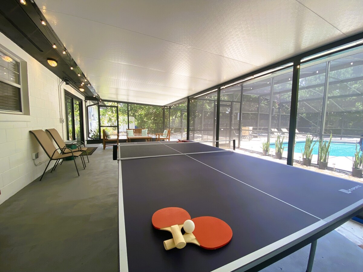 A spacious screened-in area showcases a table tennis setup, complete with paddles and balls ready for play. In the background, comfortable lounge chairs and a dining table offer additional leisure options, while the sparkling pool is visible through the screens.