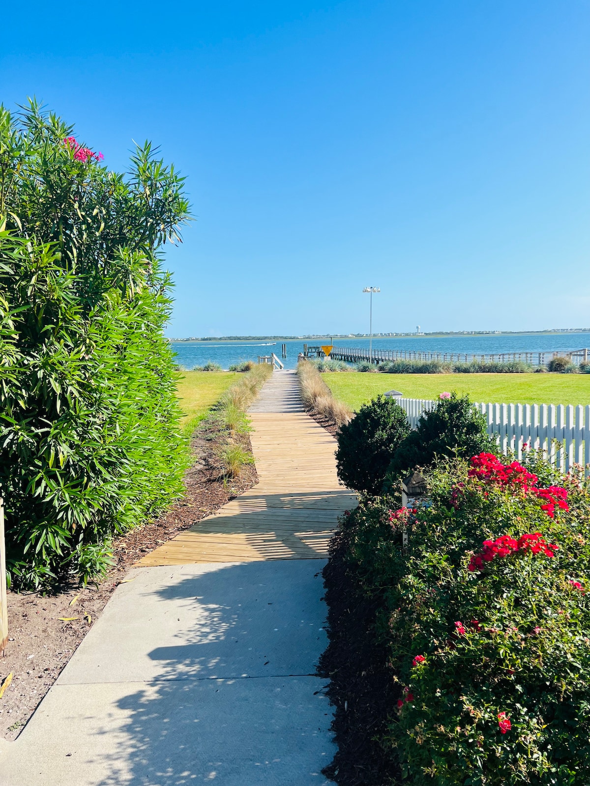 A wooden walkway leads toward the water, bordered by vibrant greenery and blooming shrubs. The pathway is flanked by a white picket fence, with views of the calm water under a clear blue sky, enhancing the inviting atmosphere of the outdoor space.