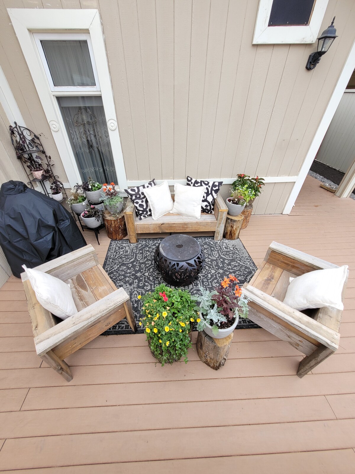 An outdoor seating area is surrounded by potted plants and flowers, creating a vibrant atmosphere. Two wooden chairs with cushions are arranged on a patterned rug, accompanied by a round table at the center. A covered barbecue grill is visible in the background.